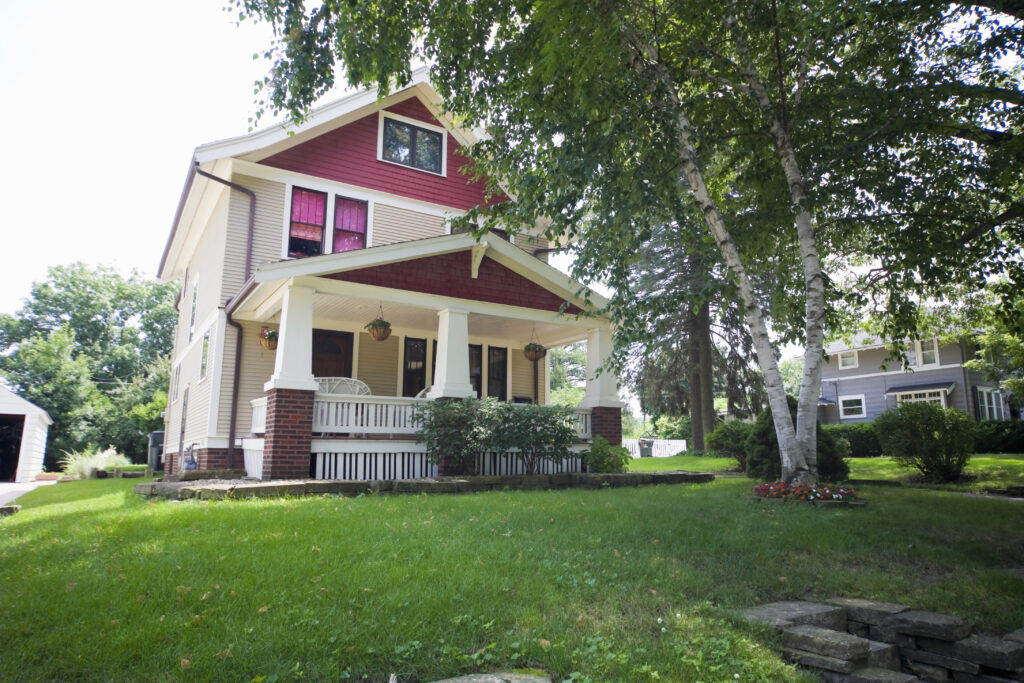 Two-story house with red siding, white trim, and a covered front porch surrounded by green lawn and trees.