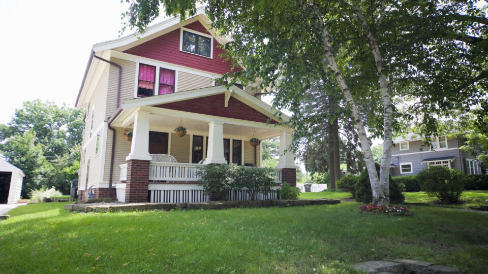 Two-story house with red siding, white trim, and a covered front porch surrounded by green lawn and trees.