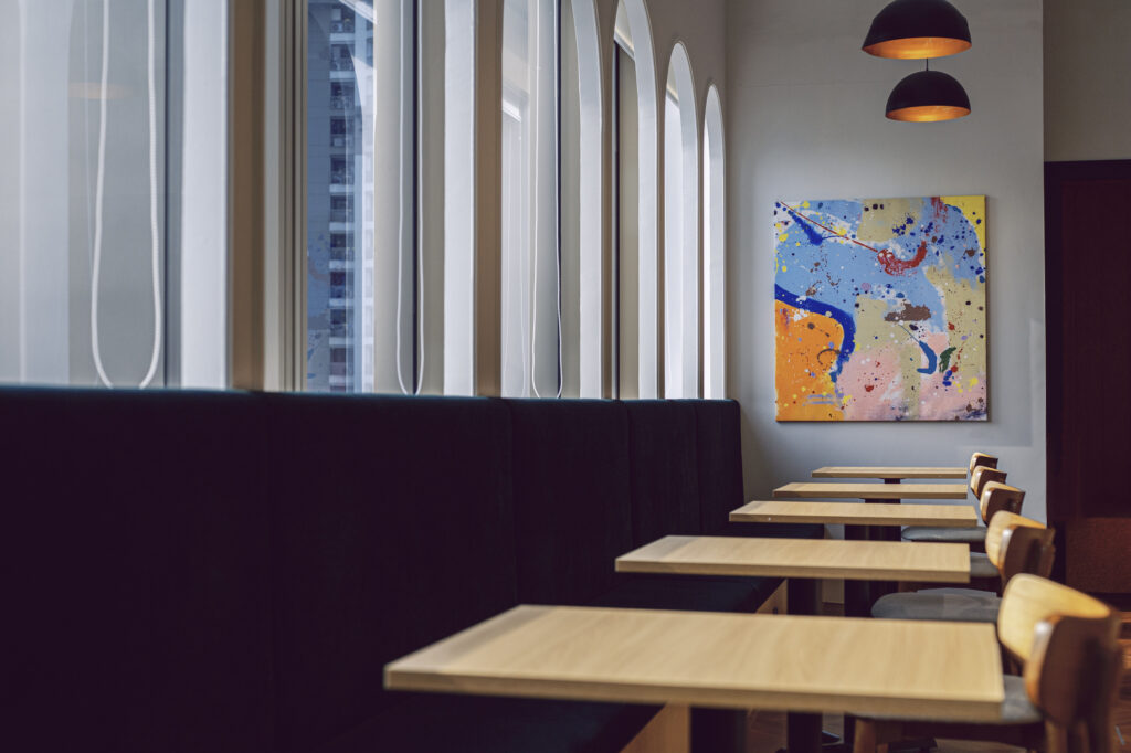 Café seating area with a long black booth, light wood tables, and a large colorful abstract painting on the wall near tall arched windows.
