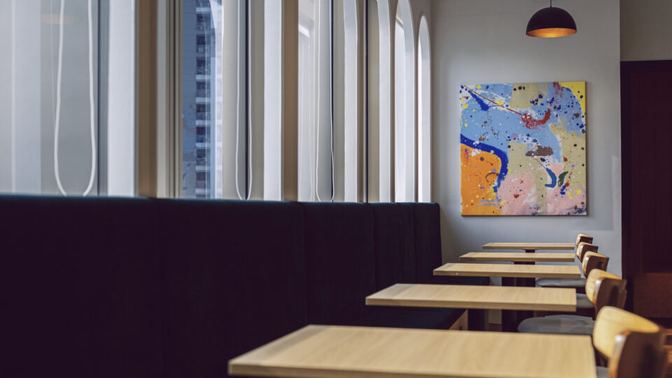 Café seating area with a long black booth, light wood tables, and a large colorful abstract painting on the wall near tall arched windows.