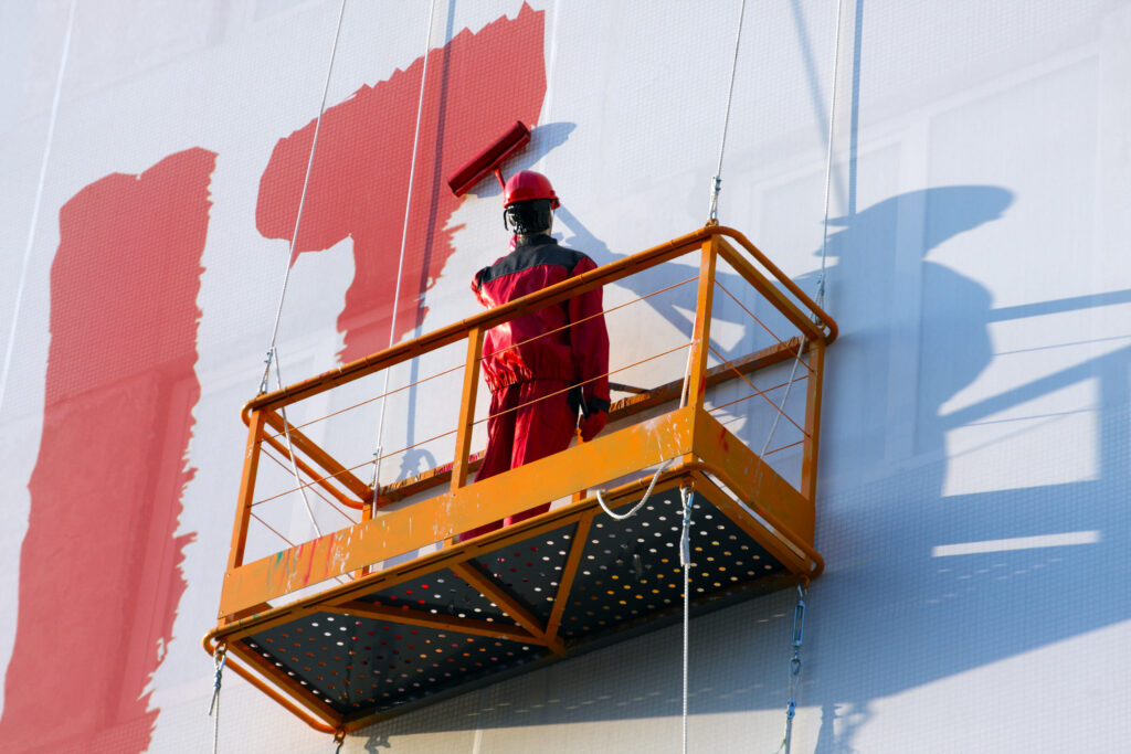 Construction worker in red protective clothing and helmet on an orange suspended platform painting a large white wall with red paint.