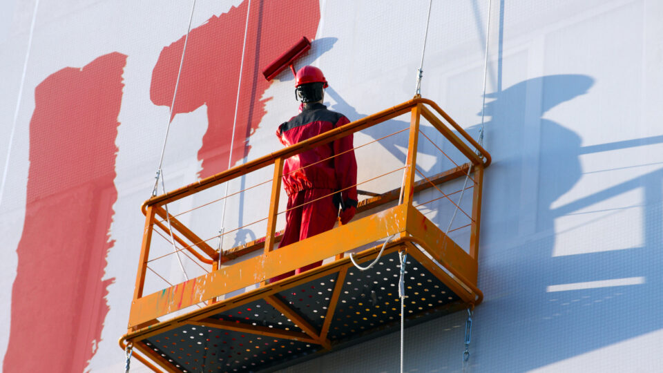 Construction worker in red protective clothing and helmet on an orange suspended platform painting a large white wall with red paint.